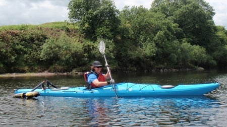 A person wearing a life jacket and sunglasses is kayaking on a calm river in a bright blue kayak. They are holding a paddle and appear to be enjoying the outdoor activity. The background features lush green trees and a grassy riverbank under a partly cloudy sky.