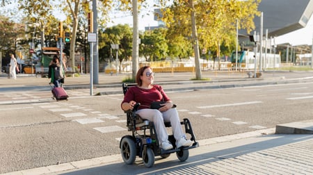A woman with glasses wearing a burgundy top and light-colored pants sits in a black power wheelchair on a paved plaza or pedestrian area. She appears relaxed and is positioned near a dropped curb where the pavement meets the street level. The setting shows an urban environment with trees lining the area, modern buildings in the background, and other pedestrians visible in the distance. The lighting suggests it's daytime with natural sunlight filtering through the tree canopy.