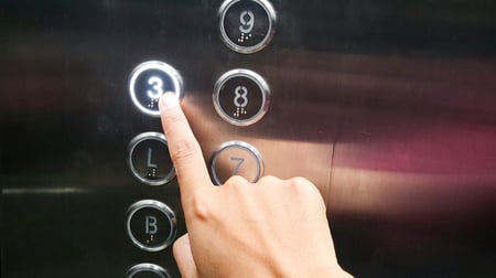 A hand with an index finger presses the illuminated number 3 button on an elevator control panel. The panel shows multiple floor buttons including basement level B, ground floor L, and floors 7, 8, and 9, all with Braille markings beneath the numbers. The button for floor 3 is lit up, indicating it has been selected.