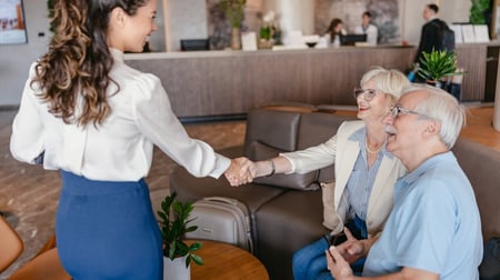 Image of a smiling young woman shaking hands in a warm friendly greeting with an elderly couple seated in a hotel lobby.
