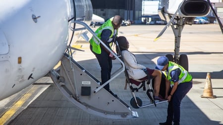 Airport ground crew members in bright green safety vests assist a passenger in a wheelchair up a mobile boarding ramp to an aircraft. Two crew members are helping guide the wheelchair user up the ramp. The scene takes place on an airport tarmac with aircraft and terminal buildings visible in the background.