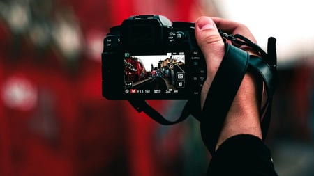 A close-up of a person holding a digital camera, with the camera screen displaying a clear image of a red street scene featuring buildings and people. The background is blurred in red tones, emphasizing the scene shown on the camera display.