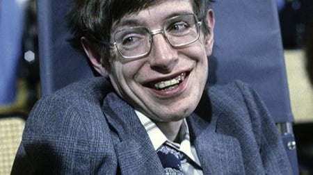  Stephen Hawking, the renowned theoretical physicist, sits in his wheelchair wearing a dark suit and tie. He has dark hair, glasses, and a warm smile, photographed in an indoor setting with blue-toned lighting in the background.