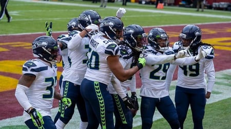 Seattle Seahawks players celebrate together on the football field, with linebacker Shaquem Griffin (#49) prominently featured among the group. The team is wearing their white away uniforms with navy blue and bright green accents, and they're gathered in the end zone area of what appears to be an opposing team's stadium with burgundy and yellow field markings.