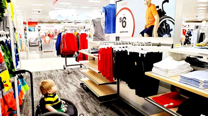 A young child with blonde hair wearing a black and yellow striped shirt sits in a wheelchair in the clothing section of a retail store. The store displays various clothing items including red and black garments on racks and shelves, with a "$6 graphic tees" price sign visible in the background. The bright, modern store environment features white shelving and displays with colorful children's clothing and accessories.